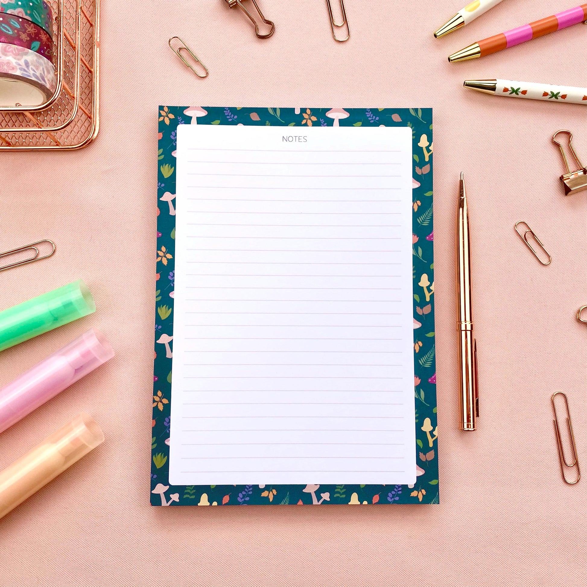 Lined notepad with a decorative blue border featuring mushrooms and leaves on a pink background with stationery items