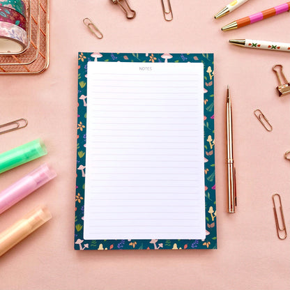 Lined notepad with a decorative blue border featuring mushrooms and leaves on a pink background with stationery items