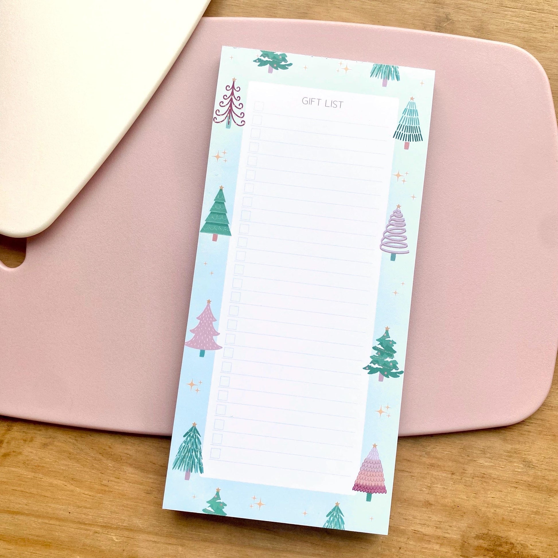 Green gift list notepad with Christmas tree design on a pink surface