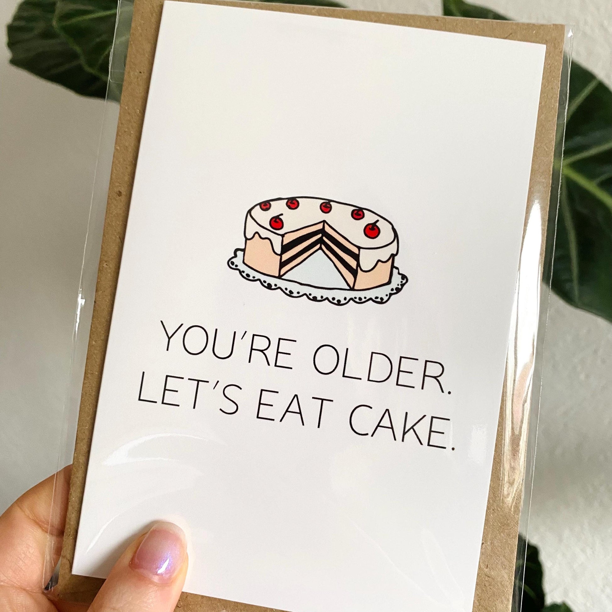 Greeting card with a cake illustration and text 'You're older. Let's eat cake.' held by a hand.