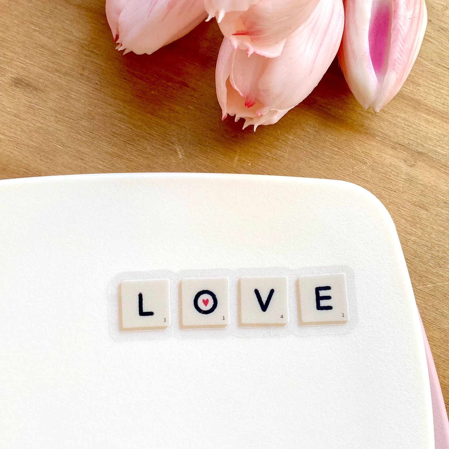 A sticker with Scrabble tiles spelling 'LOVE' on a white surface with pink tulips in the background.