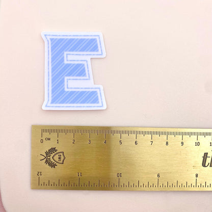 Blue striped letter 'E' sticker with a ruler for scale on a beige background