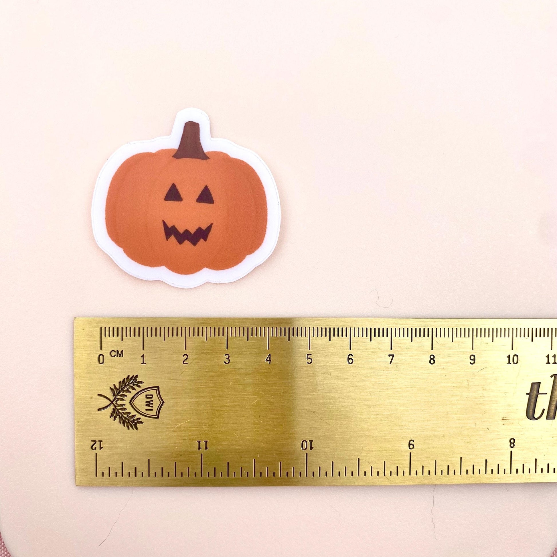 Orange jack-o-lantern pumpkin sticker against a pink background with a gold ruler for scale.