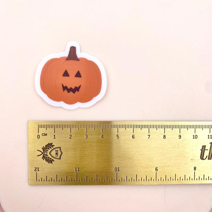 Orange jack-o-lantern pumpkin sticker against a pink background with a gold ruler for scale.