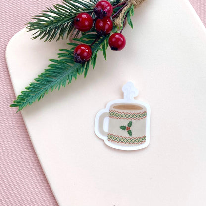 Decorative sticker of a mug with holly design on a white surface with a pink border.