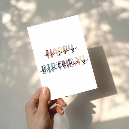 Hand holding a white card with 'Happy Birthday' text in rainbow colours against a light background