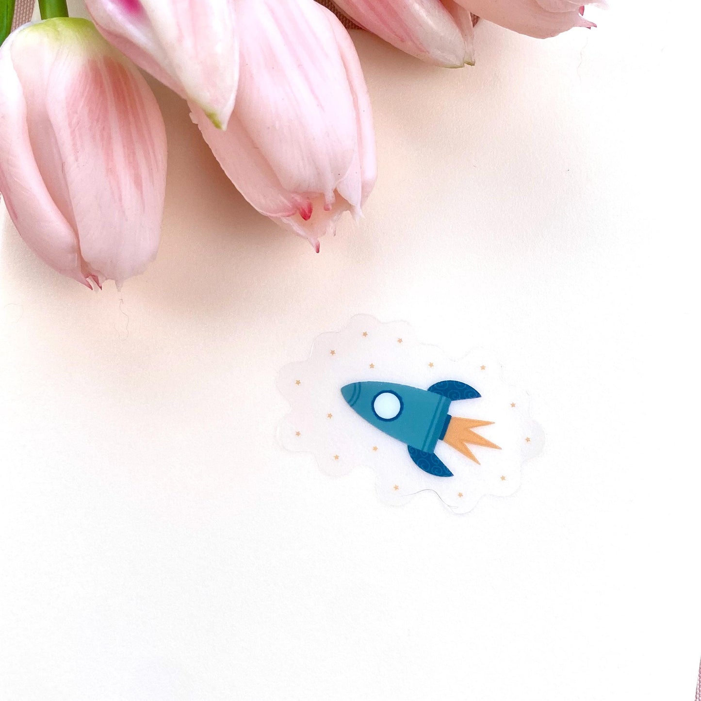 Transparent sticker with teal blue rocket against a cream background with pink tulips.