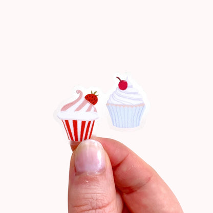 A sticker with two tiny cupcakes in pink and blue held by hand against a cream background