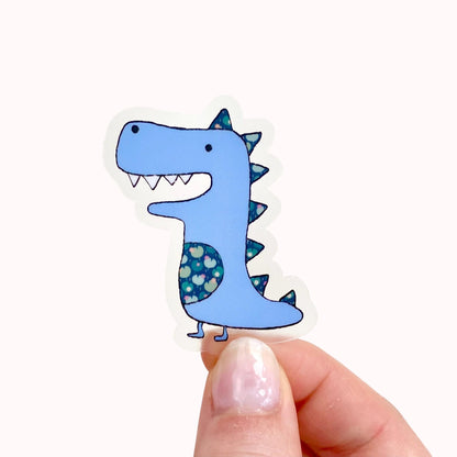 Cartoon dinosaur sticker held between fingers on a white background