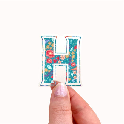 Colourful letter 'H' sticker with floral design held by a hand on a white background