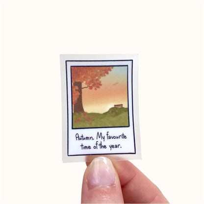 Sticker with a miniature polaroid with an illustration of an autumn scene with text held by a hand on a white background
