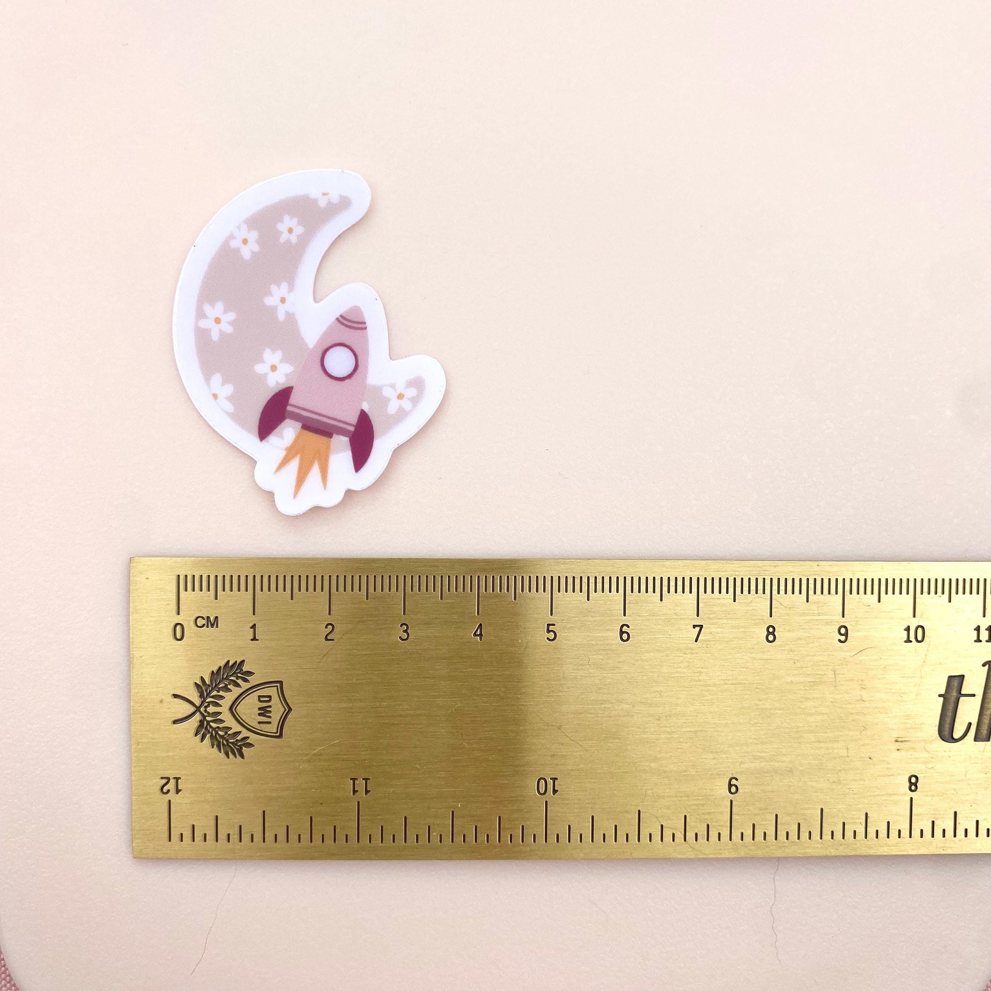 Decorative sticker with pink rocket and floral moon design on a white background with a gold ruler for scale.