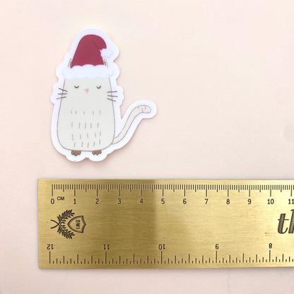 Sticker of a cat wearing a Santa hat next to a ruler on a beige background