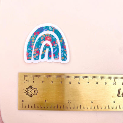 Decorative rainbow sticker with floral patterns on a white background with a gold ruler for scale.