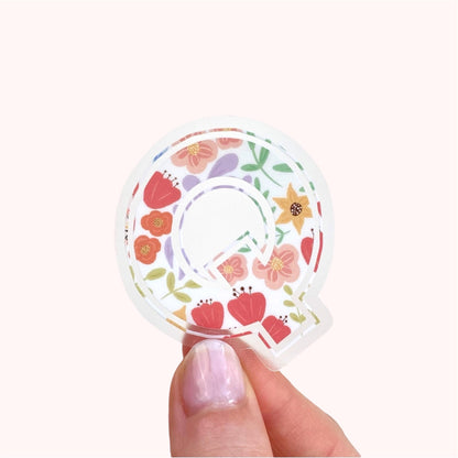 Decorative letter 'Q' sticker with floral patterns held between fingers against a light background.