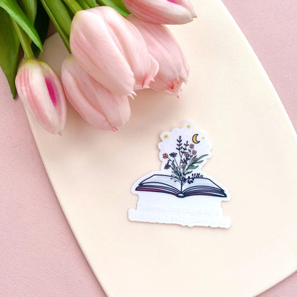 Sticker of an open book with flowers and quote on a pink surface with tulips
