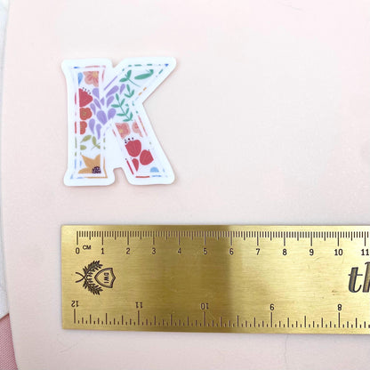 Floral letter 'K' sticker next to a ruler on a pink background