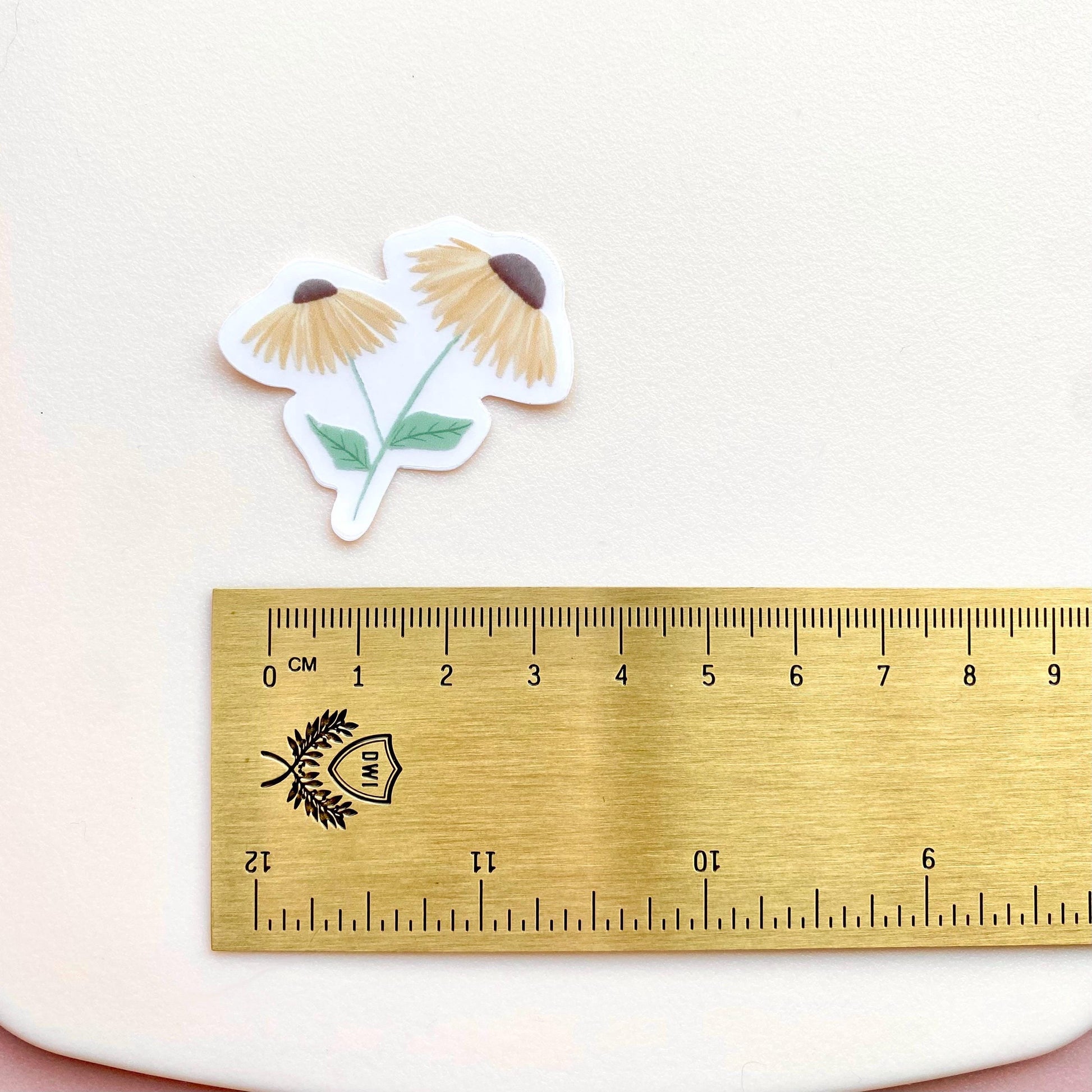 A sticker with 2 yellow daisies against a cream background with a gold ruler for scale.