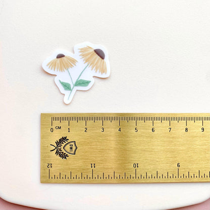 A sticker with 2 yellow daisies against a cream background with a gold ruler for scale.
