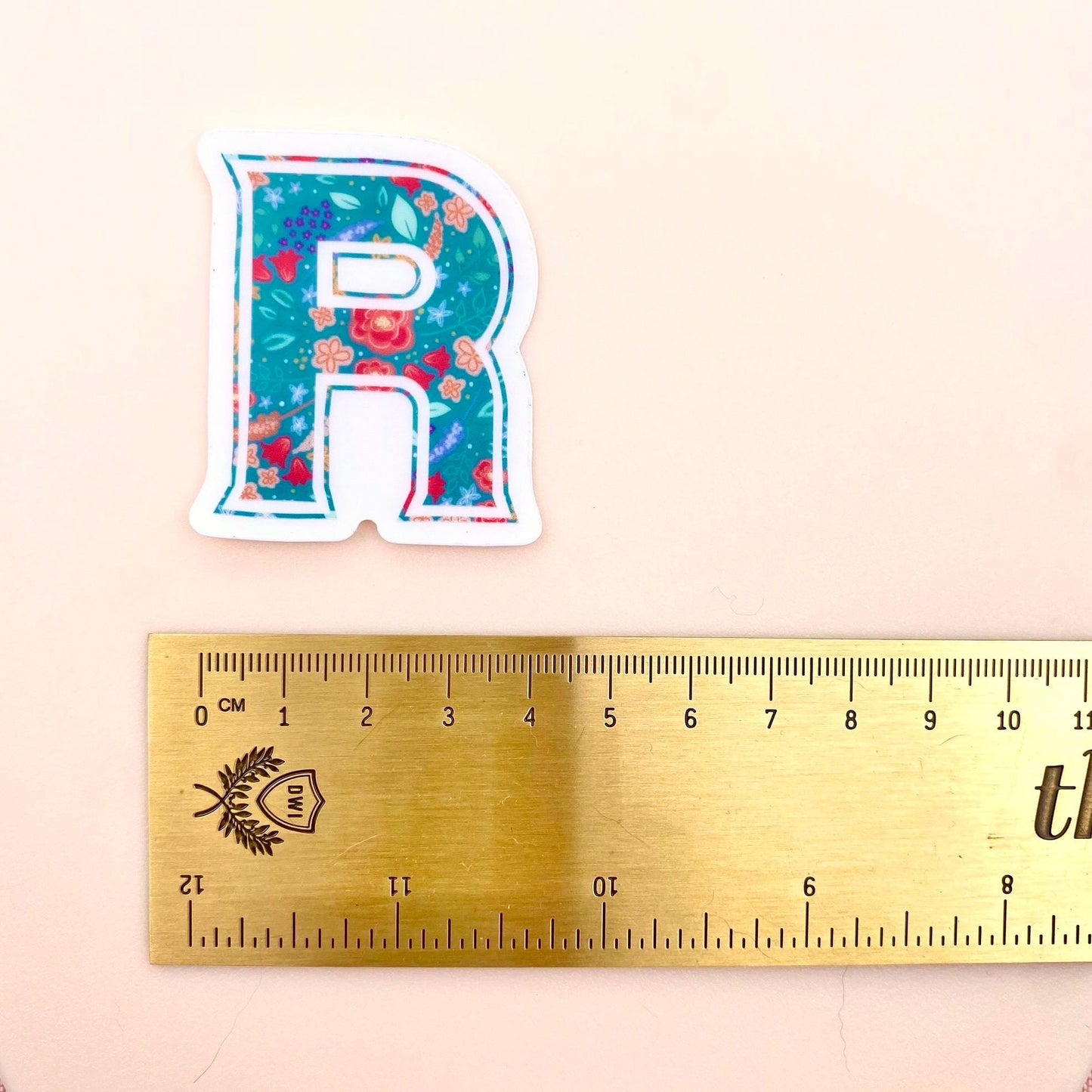 Sticker with a floral design on a letter 'R' next to a ruler for scale