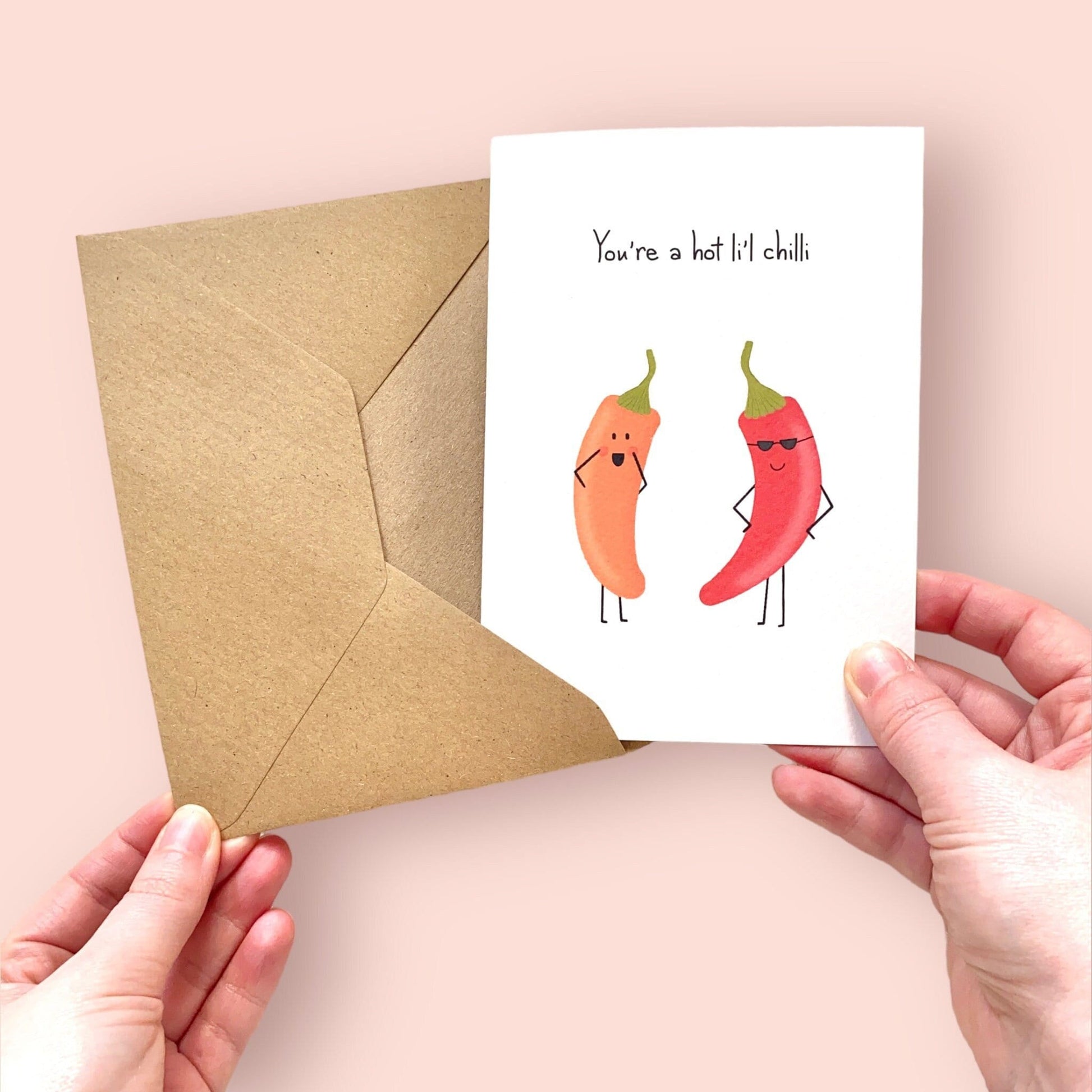 Card with cartoon red and orange  peppers and 'You're a hot li'l chilli' text and an envelope held by hands on a pink background
