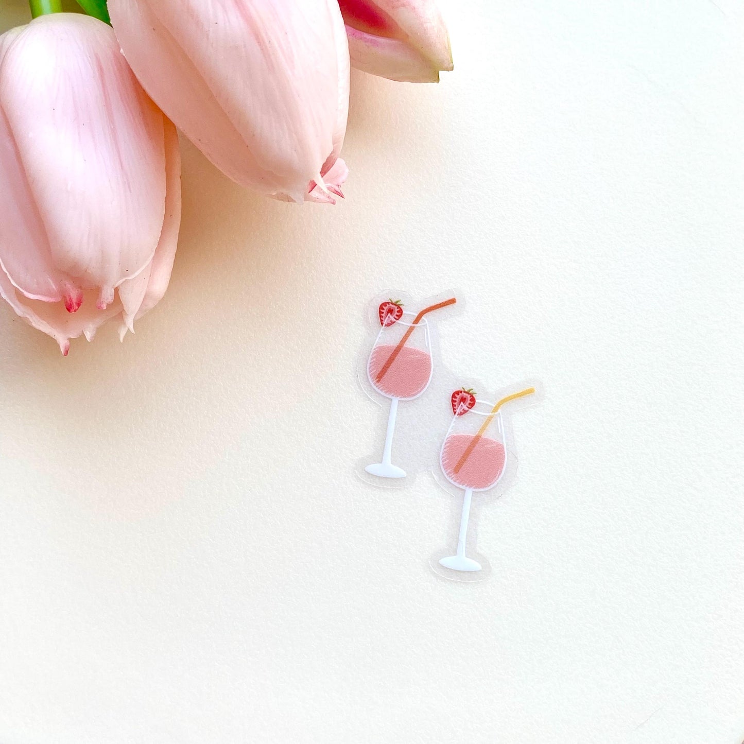 A sticker of two pink drinks in wine glasses with colourful straws and strawberries on a white surface.