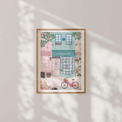 Framed illustration of a bookshop with a pink bicycle outside on a light grey wall.
