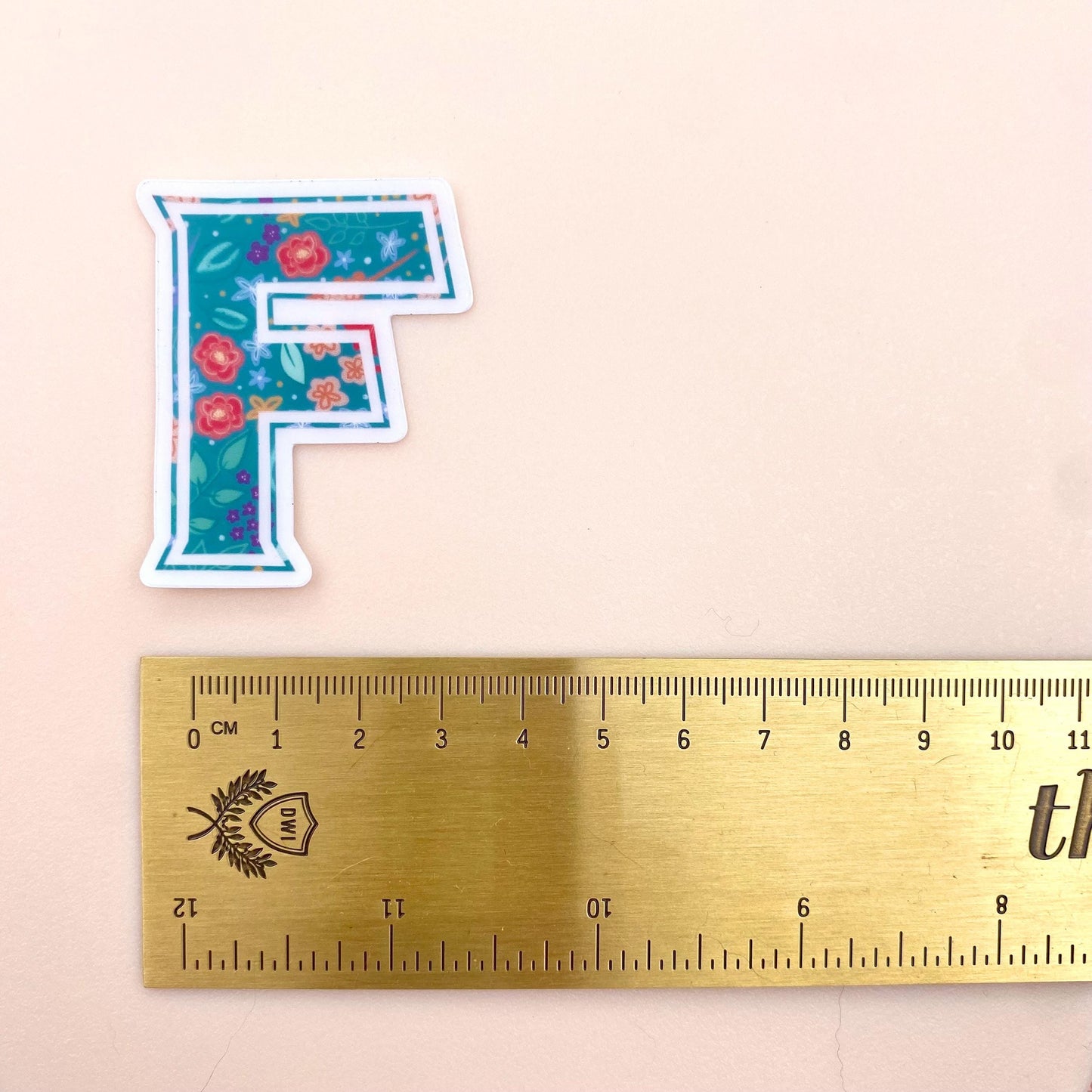 Floral letter 'F' sticker next to a ruler on a beige background