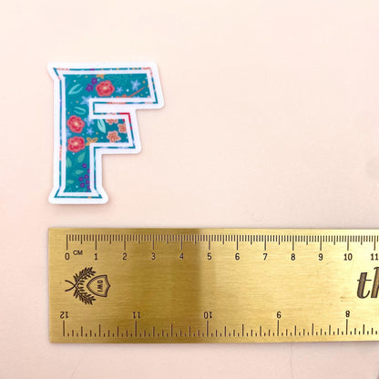 Floral letter 'F' sticker next to a ruler on a beige background