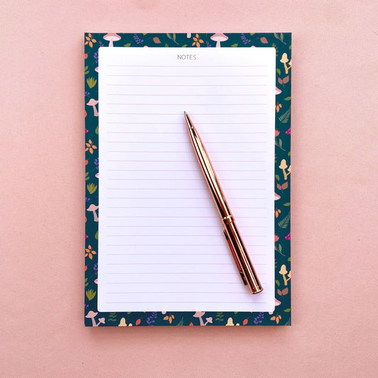 Lined notepad with a decorative blue border featuring mushrooms and leaves on a pink background with a rose gold pen