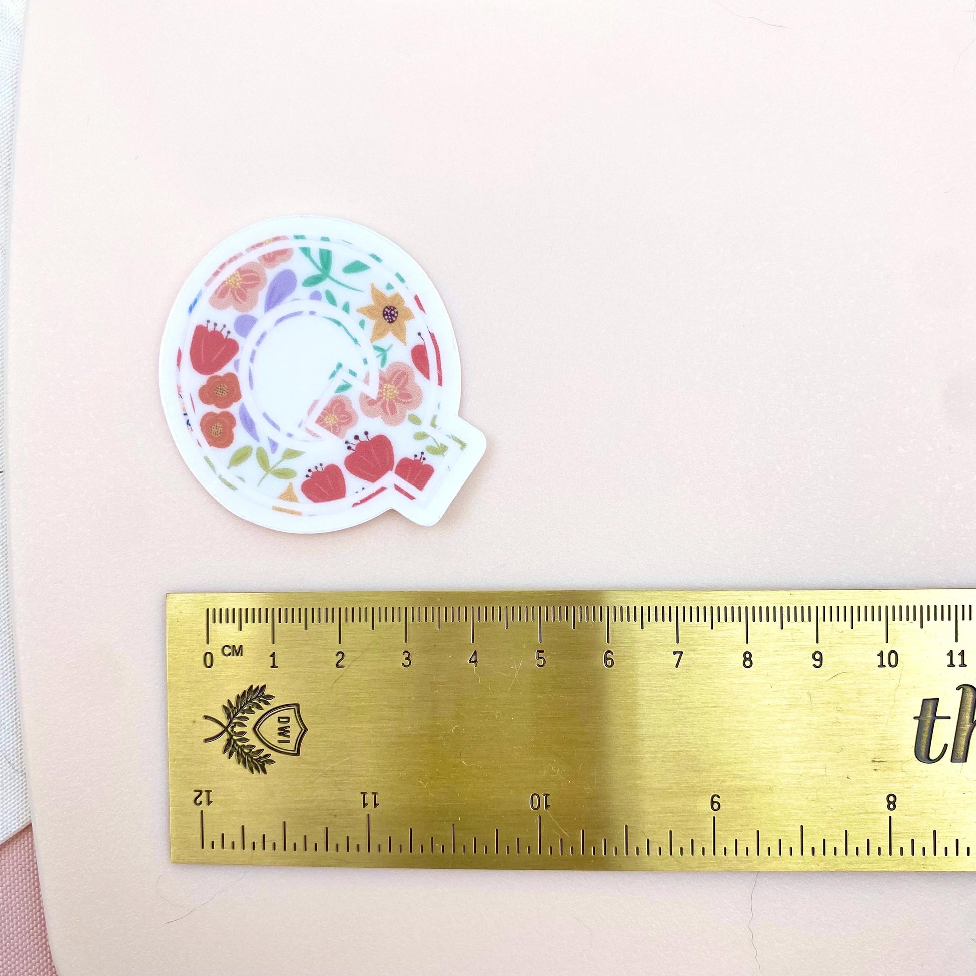 Decorative letter 'Q' sticker with floral patterns on a light background with a gold ruler for scale.