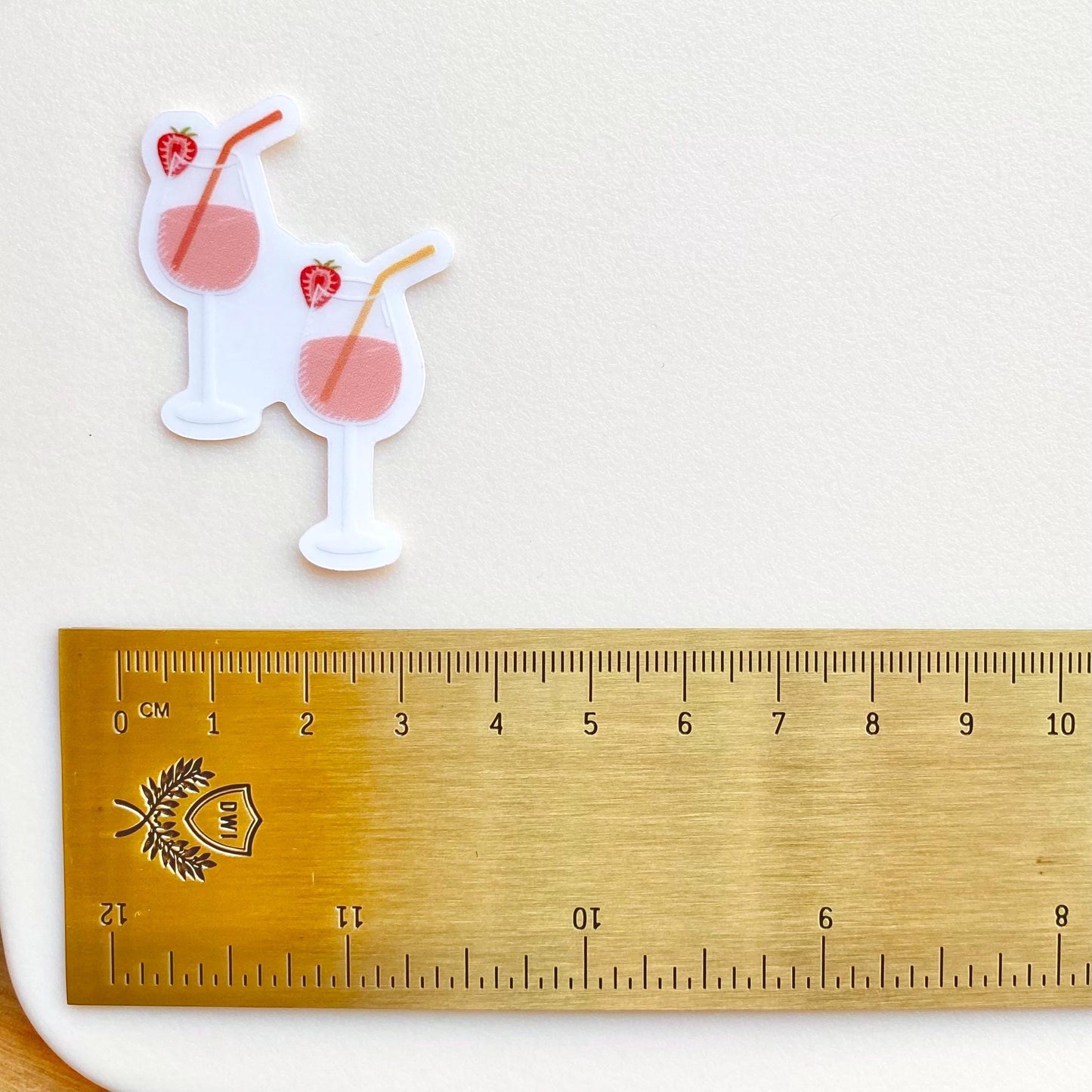 A sticker of two pink drinks in wine glasses with colourful straws and strawberries on a white surface a gold ruler for scale.