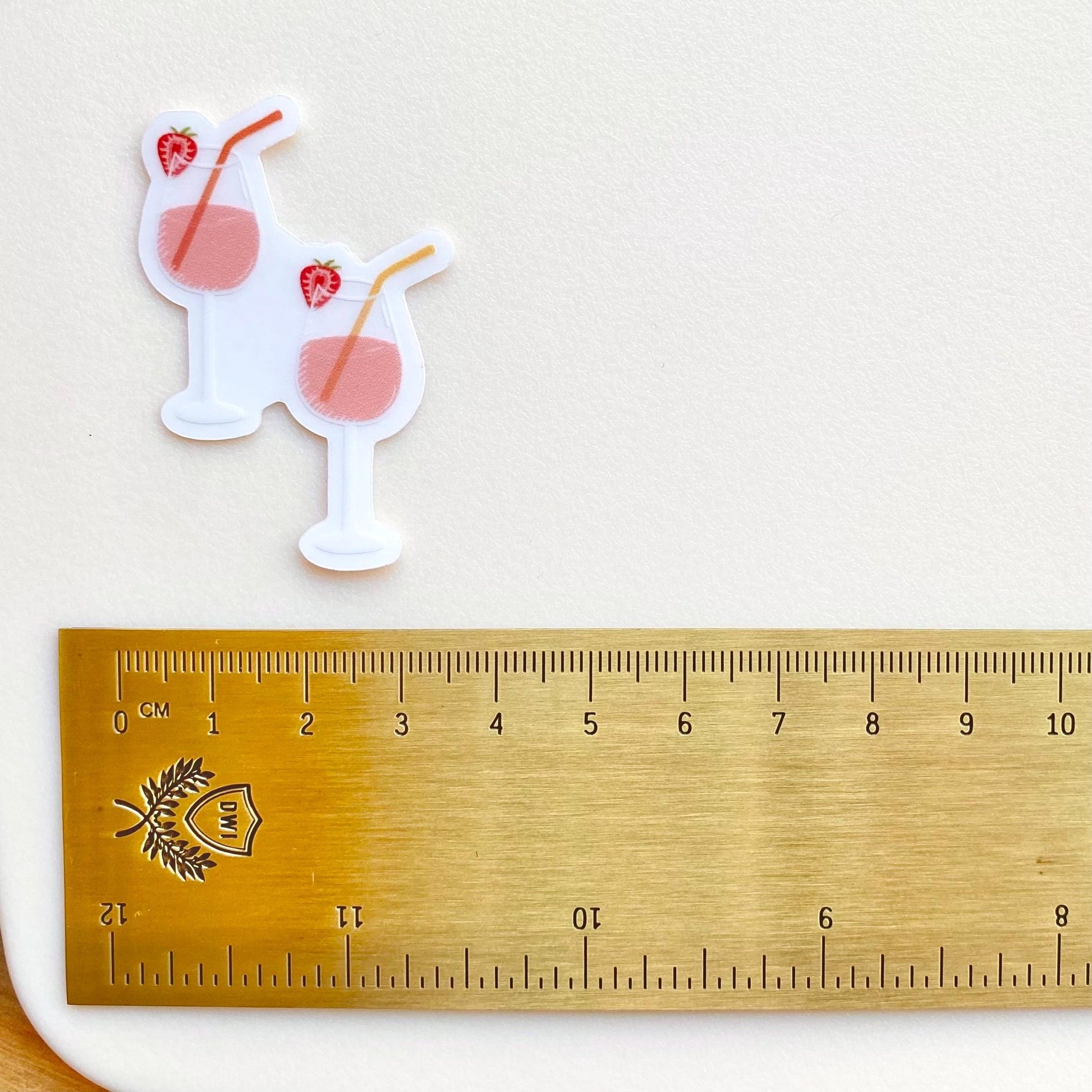 A sticker of two pink drinks in wine glasses with colourful straws and strawberries on a white surface a gold ruler for scale.