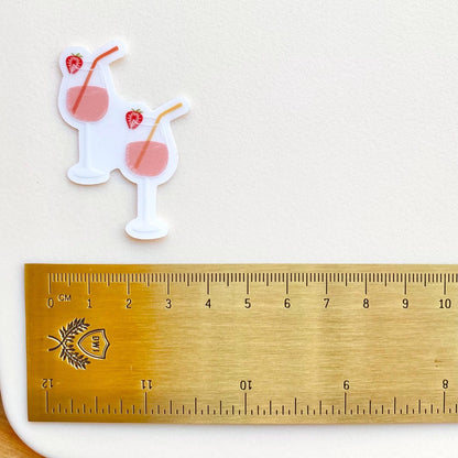 A sticker of two pink drinks in wine glasses with colourful straws and strawberries on a white surface a gold ruler for scale.