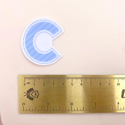 Blue striped  letter 'C' transparent sticker on a white background with a gold ruler for scale
