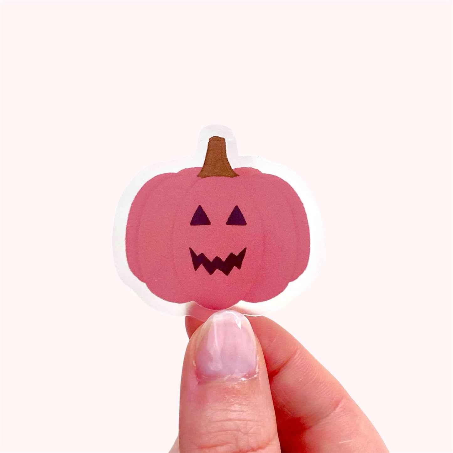 Pink pumpkin sticker with a carved face held by a hand on a white background