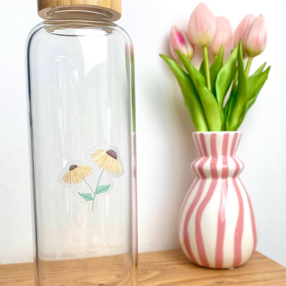A sticker with 2 yellow daisies against a glass bottle besides a pink striped vase of tulips.