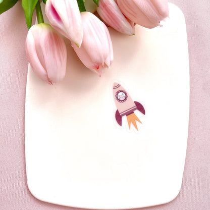 A pink rocket sticker with a grey cat in the window against a cream background with pink tulips