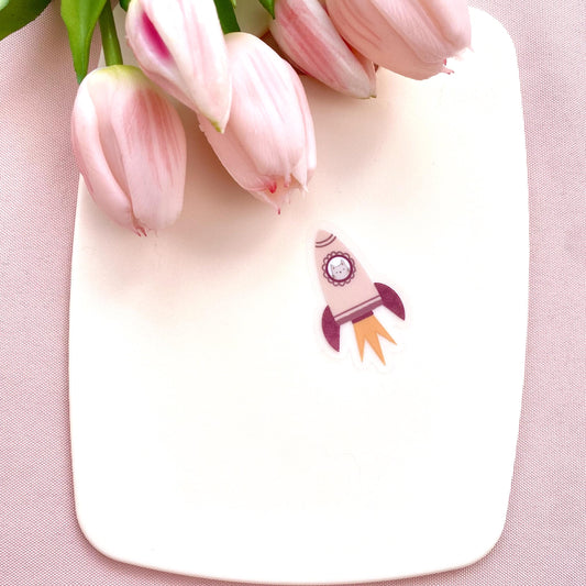 A pink rocket sticker with a grey cat in the window against a cream background with pink tulips