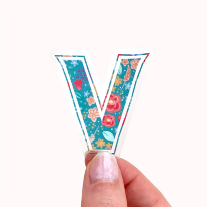 Hand holding a colourful letter 'V' sticker with floral design on a white background