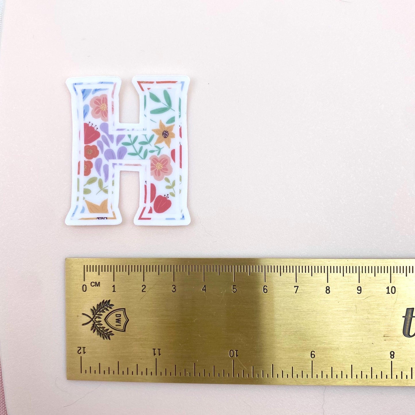 Letter 'H' sticker with floral design on a white background with a gold ruler for scale.