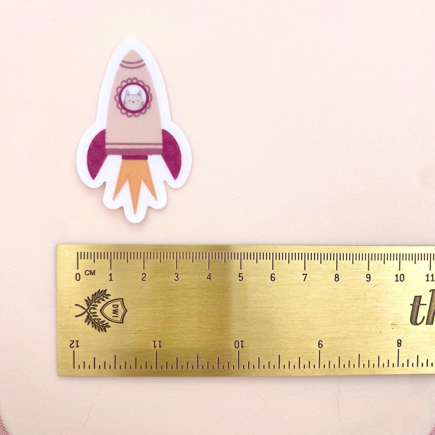 A pink rocket sticker with a grey cat in the window against a cream background with a gold ruler for scale.