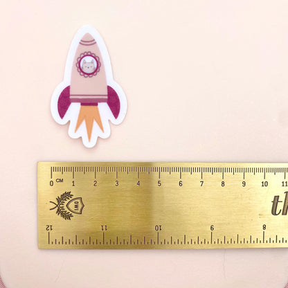 A pink rocket sticker with a grey cat in the window against a cream background with a gold ruler for scale.