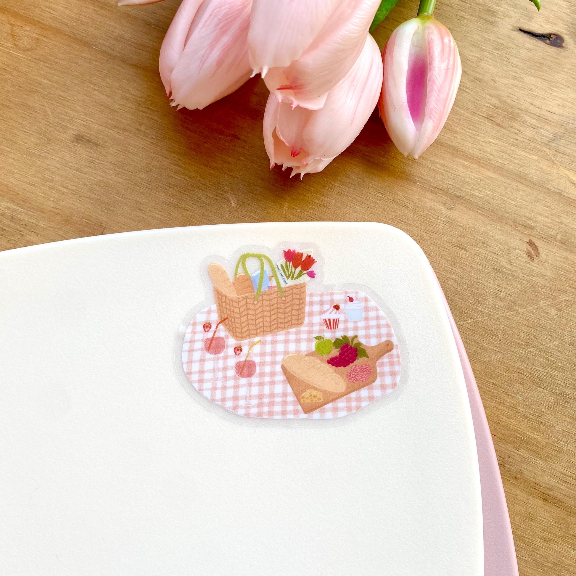 Sticker of a picnic scene on a white surface with pink tulips in the background.
