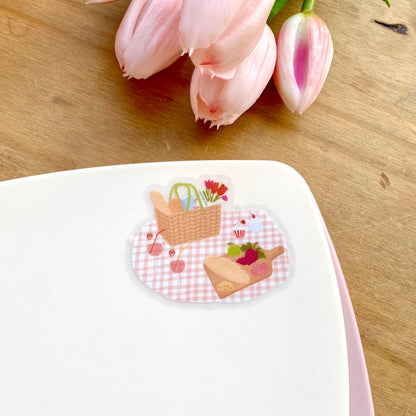 Sticker of a picnic scene on a white surface with pink tulips in the background.