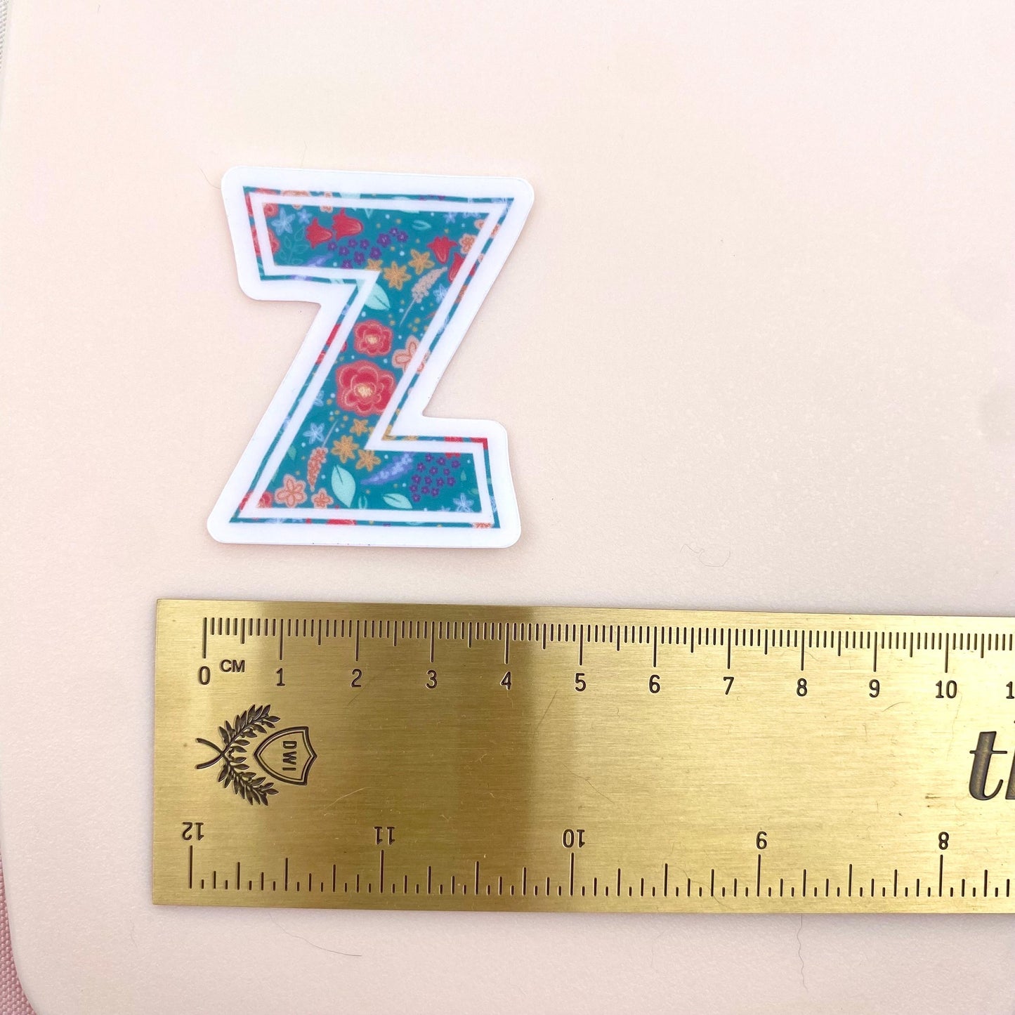 Floral letter 'Z' sticker next to a ruler on a light pink background