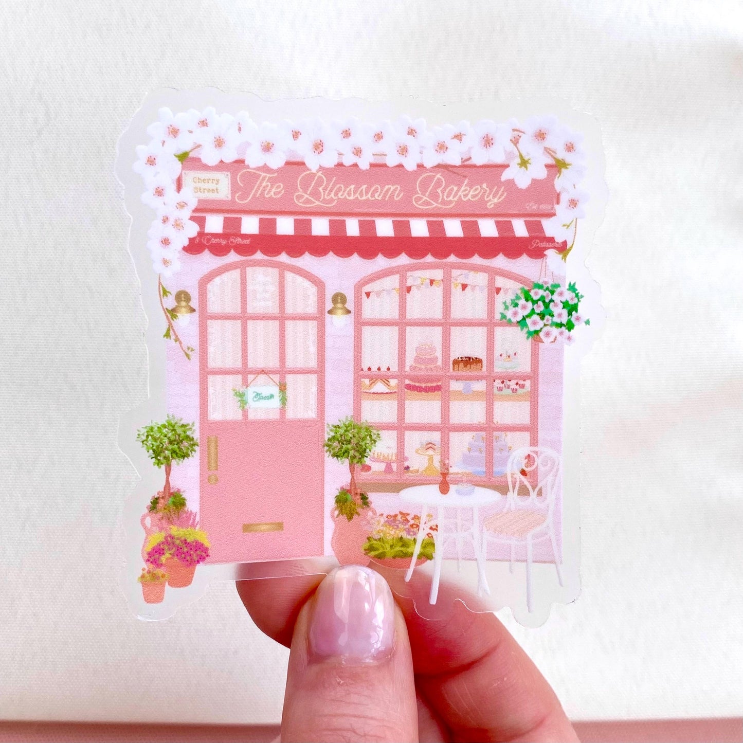 Sticker of 'The Blossom Bakery' with a pink building design held by a hand.