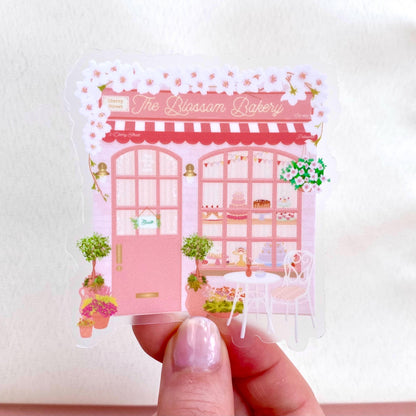 Sticker of 'The Blossom Bakery' with a pink building design held by a hand.
