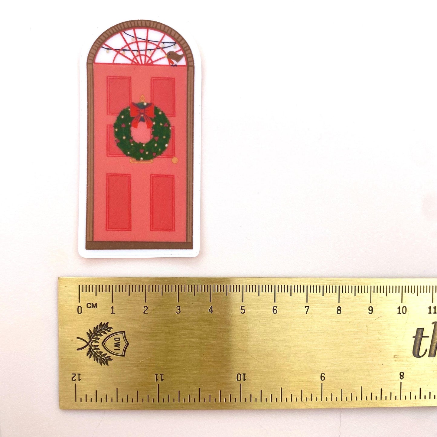 Sticker with a red door and wreath design next to a ruler for scale on a white background