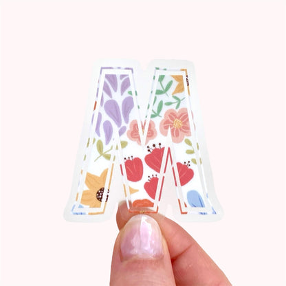 Decorative letter 'M' sticker with floral patterns held by a hand on a white background